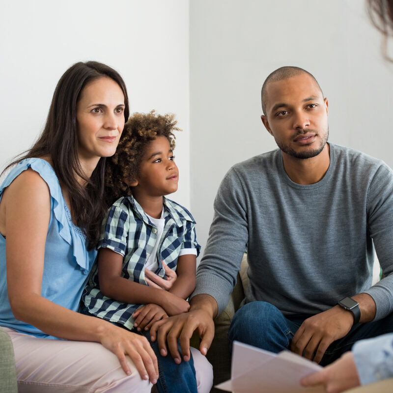 Serious couple with son in a deep conversation with family counselor. Multiethnic parents talking about adoption with their counselor. Family at the financial planning consultation.